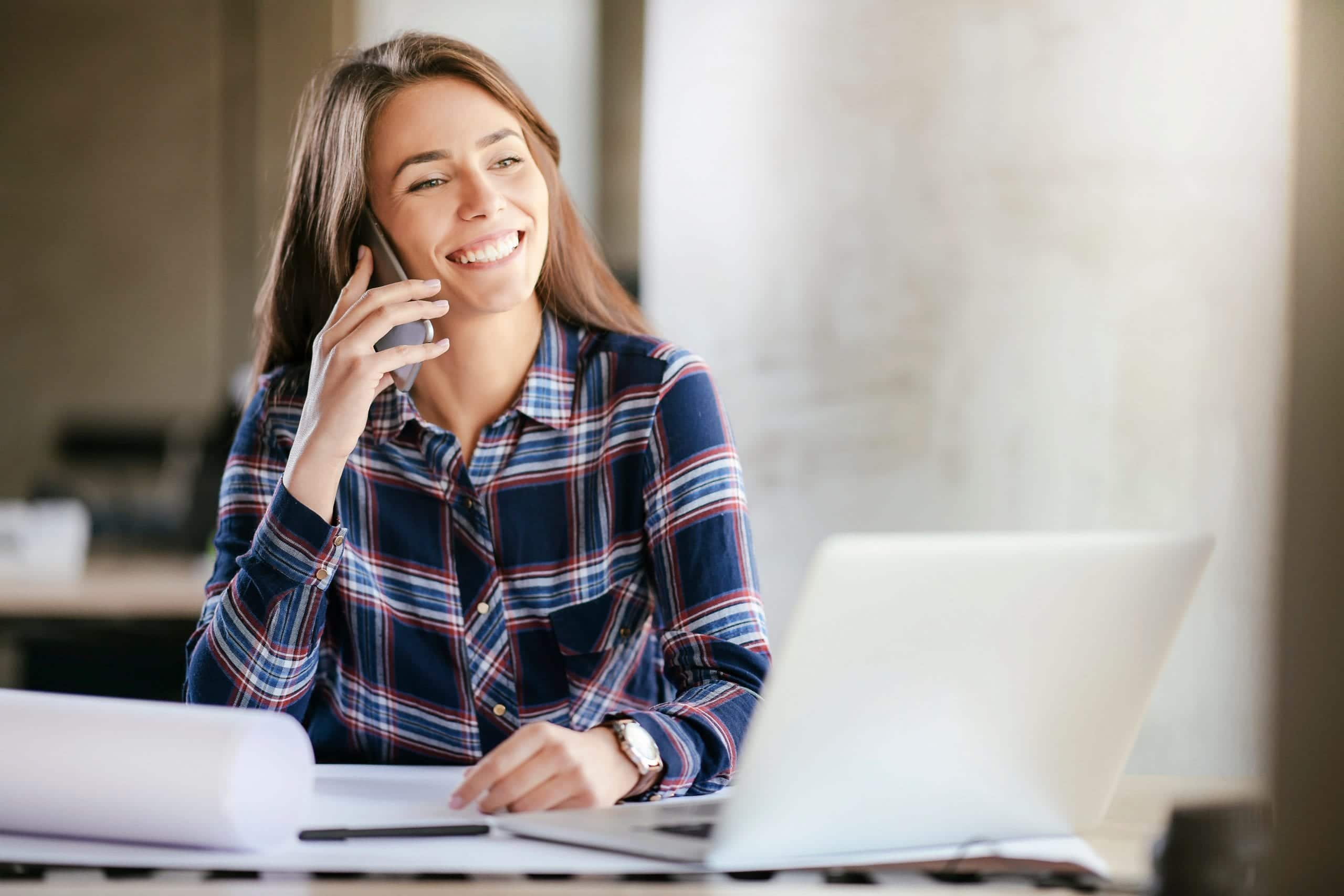 young woman seated at desk in front of laptop while talking on phone and smiling