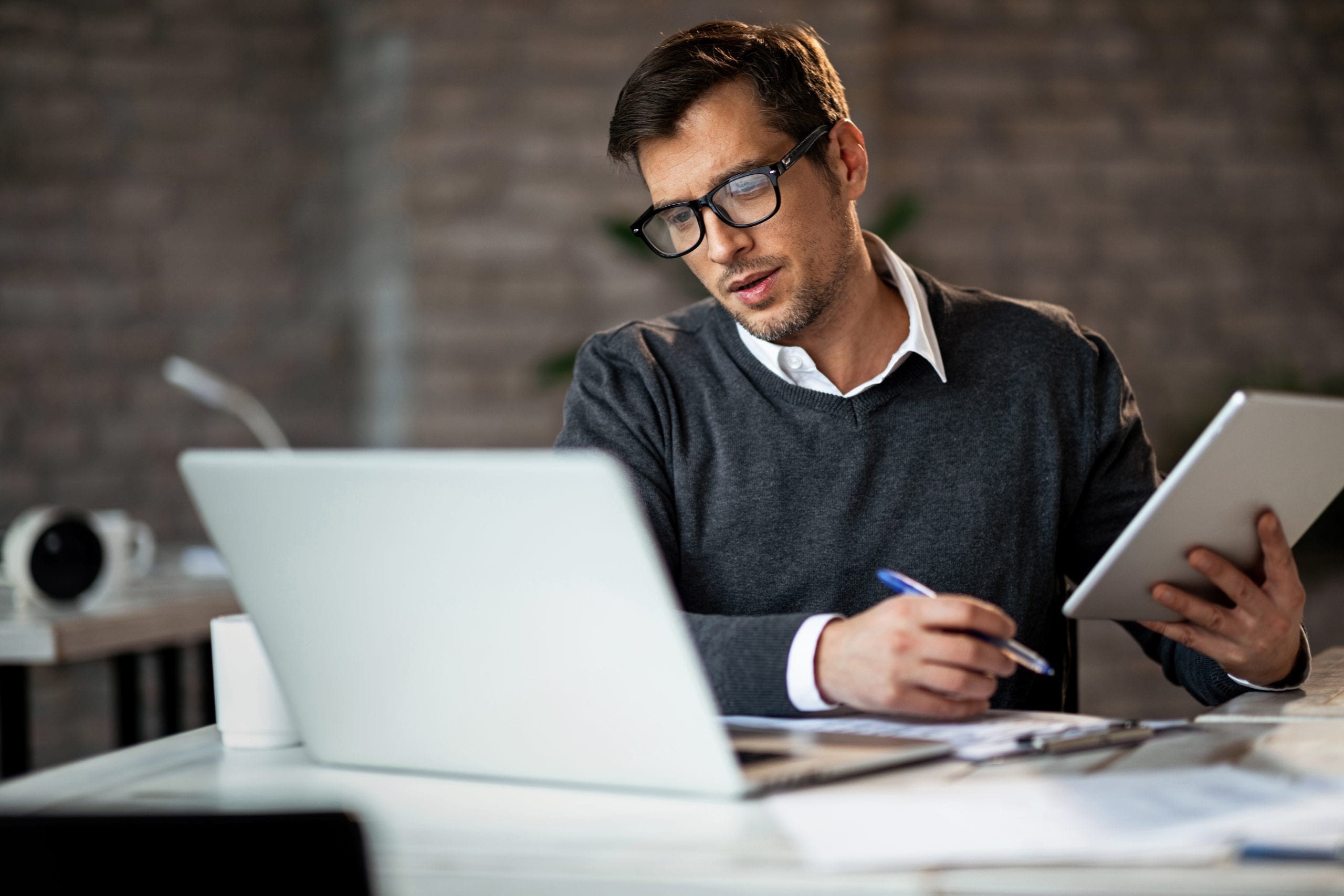 young man in office setting working while holding tablet and looking at laptop