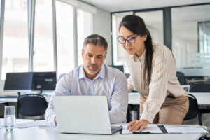 a person looks over the shoulder of another while looking at a laptop and explaining the differences between medical billing vs. revenue cycle management