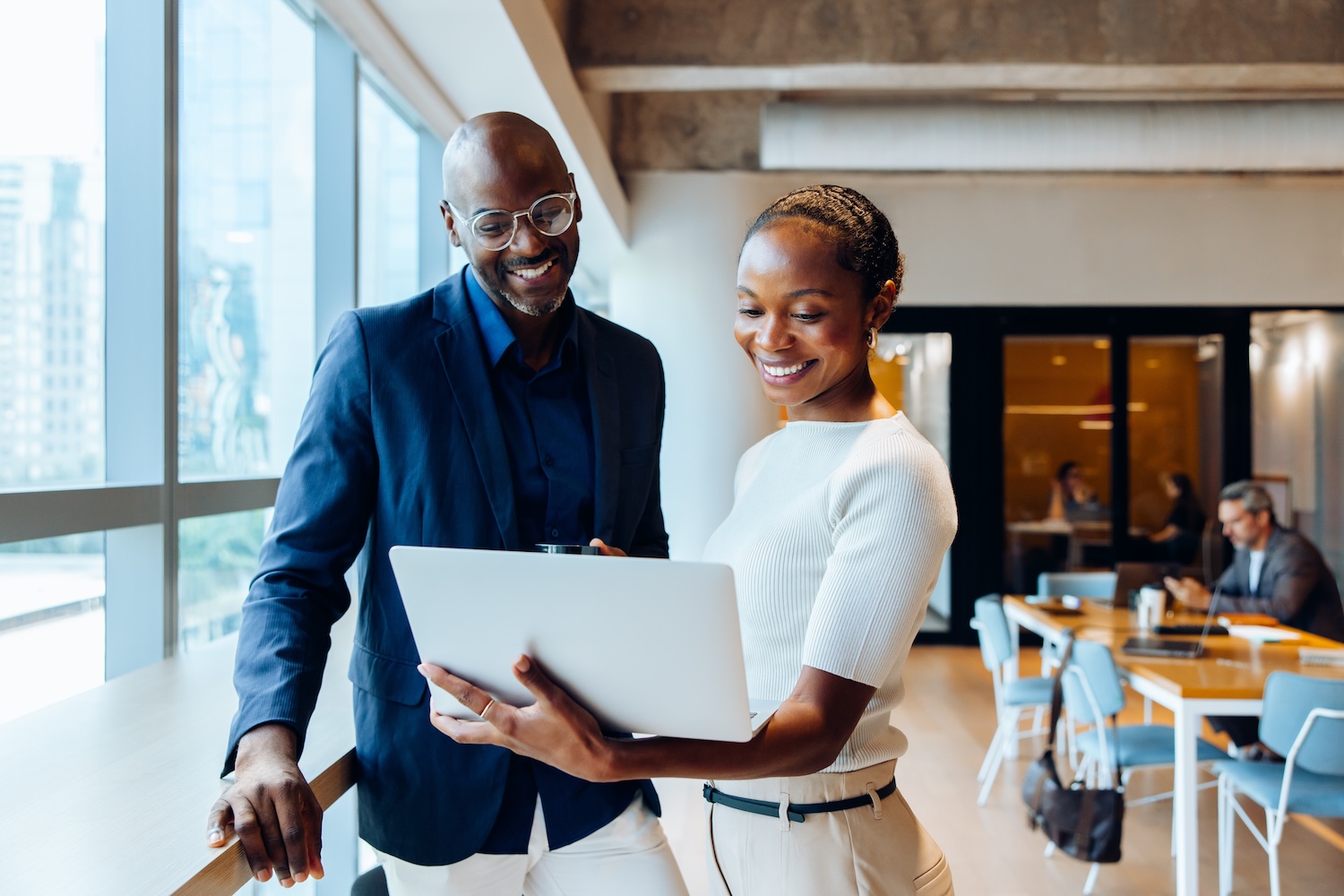 Two smiling professionals standing by a large window in a modern office, looking at a laptop together and discussing work.