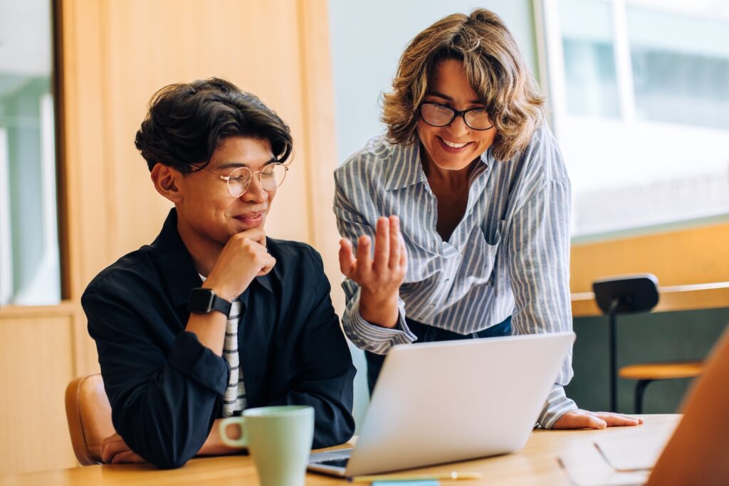 Two colleagues collaborating at a desk, smiling as they discuss something on a laptop screen in a bright, modern office.