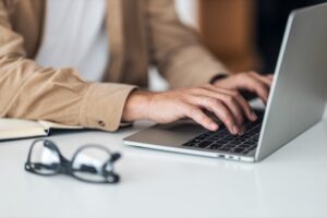 Close-up of a person typing on a laptop with eyeglasses on the table.